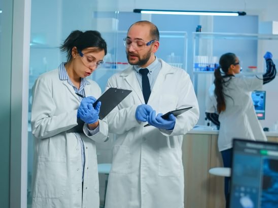Group of medical researchers discussing about vaccine development standing in equipped lab pointing on tablet and taking notes. Doctors examining virus evolution using high tech researching diagnosis